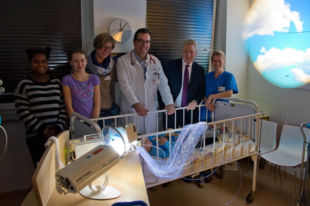 Ärzte, Therapeuten und Patienten stehen an einem Patientenbett und betrachten den Snoezelenwagen.